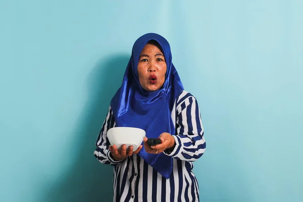 Zszokowana Azjatka Średnim Wieku Niebieskim Hidżabie Ogląda Telewizję Kierując Pilota — Zdjęcie stockowe
