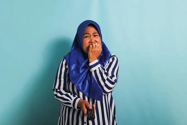 A worried middle-aged Asian woman in a blue hijab and a striped shirt is biting her fingers while holding a remote control and watching a horror movie on TV. She is isolated on a blue background