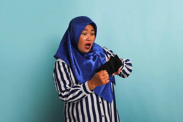 青いヒジャーブとストライプのシャツを着た驚くべき中高年アジアの女性がショックを受けた驚きの表情で彼女の空の財布を調べています 彼女は青い背景に隔離されている — ストック写真