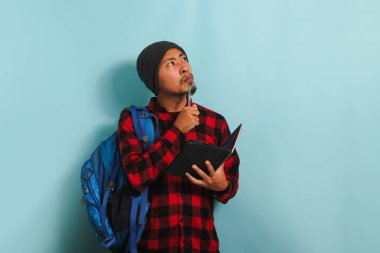 Sırt çantası takmış, bereli ve kırmızı flanel gömlekli düşünceli genç Asyalı bir öğrenci derin bir düşünceye dalmış elinde kalemle fotokopi odasına bakarken mavi bir arkaplana bakıyor.