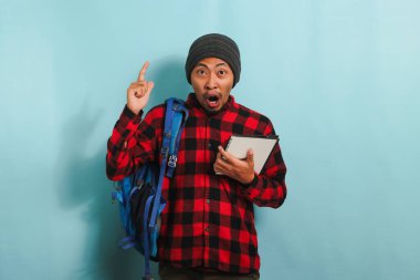 Sırt çantası takmış, bereli ve kırmızı flanel gömlekli heyecanlı bir Asyalı genç öğrenci mavi arka planda dururken fotokopi alanını işaret ediyor.