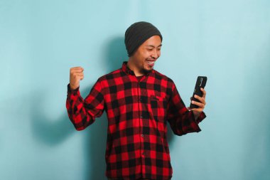 Heyecanlı, bereli, kırmızı flanel gömlekli Asyalı genç adam elinde iyi haberlerle akıllı telefonu tutarken EVET jesti yapıyor.