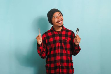 Şapkalı, kırmızı flanel gömlekli, elinde çatal ve kaşık tutan Asyalı genç adam, mavi bir arka planda izole edilmiş, mutlu ve yemek için istekli görünüyor.