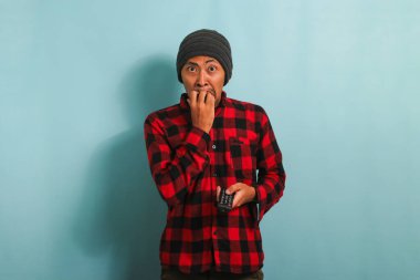 Şaşkın Asyalı şapkalı ve kırmızı gömlekli genç bir adam TV 'de şok edici haberler izliyor, mavi arka planda izole edilmiş.