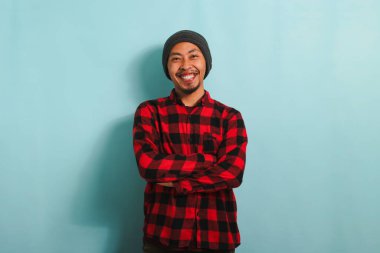 Şapkalı, kırmızı flanel gömlekli, kendine güvenen Asyalı genç bir adam kollarını kavuşturmuş kameraya bakıyor. Mavi bir arka planda izole edilmiş.