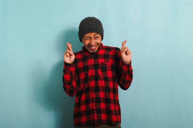 Bere şapkalı, kırmızı ekose gömlekli heyecanlı bir Asyalı genç adam mavi arka planda dikilirken rüyalarının gerçek olmasını umarak parmaklarını çapraz yapıp seviniyor.