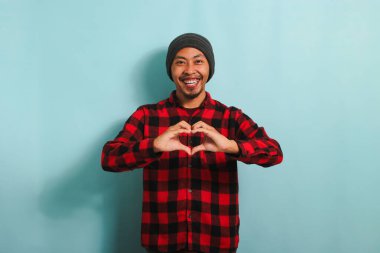 Şapkalı, kırmızı flanel gömlekli gülümseyen Asyalı genç adam mavi arka planda dururken bir aşk jesti gösteriyor.