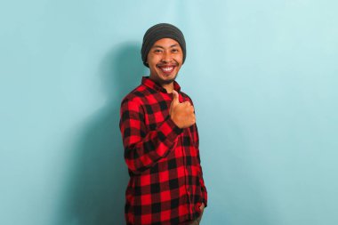 Şapkalı, kırmızı ekose gömlekli, kendine güvenen Asyalı genç adam iyi bir jest yapıyor ve kendini mavi arka planda yalnız hissediyor.