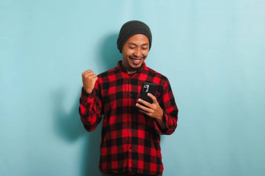 Heyecanlı, bereli, kırmızı flanel gömlekli Asyalı genç adam elinde iyi haberlerle akıllı telefonu tutarken EVET jesti yapıyor.