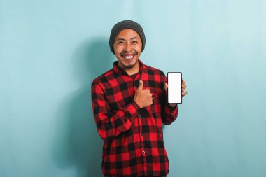 Şapkalı, kırmızı flanel gömlekli neşeli Asyalı genç bir adam boş bir ekran akıllı telefonu gösteriyor, başparmak işareti yapıyor ve mavi arka planda dururken yeni bir uygulama öneriyor.