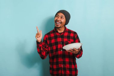 Heyecanlı, bereli ve kırmızı gömlekli Asyalı genç adam boş fotokopi odasını gösterirken elinde mavi arka planda izole edilmiş beyaz bir tabak tutuyor.