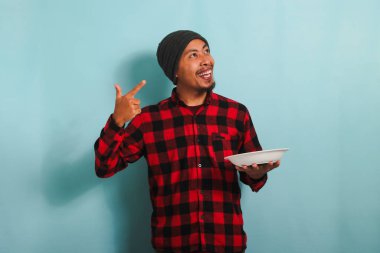 Bereli ve kırmızı flanel gömlekli heyecanlı Asyalı genç adam boş fotokopi alanını gösterirken elinde mavi arka planda izole edilmiş beyaz bir tabak tutuyor.