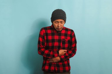 Bere şapkalı, kırmızı ekose gömlekli mutsuz bir Asyalı adam karın ağrısından muzdarip, eğiliyor ve ellerini karnının üstüne koyup mavi arka planda duruyor.