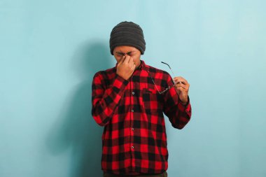 Çok çalışan, bereli, kırmızı flanel gömlekli, gözlüklü, kuru gözlerinde ağrı hisseden bilgisayar üzerinde saatlerce çalıştıktan sonra mavi arka planda izole edilmiş, baş ağrısı çeken Asyalı genç bir adam.