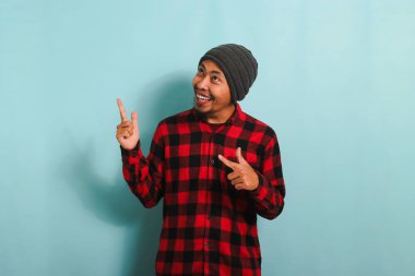 Heyecanlı, bereli ve kırmızı flanel gömlekli Asyalı genç adam parmağını fotokopi alanının solunda mavi arka planda izole edilmiş bir şekilde gösteriyor.