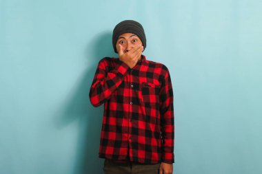 Şapkalı ve kırmızı flanel gömlekli genç Asyalı adam ağzını kapatıyor. Bu da demek oluyor ki, çenesini kapatmaya zorlanıyor. Mavi arka planda tek başına.