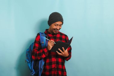 Sırt çantası takan, bereli ve kırmızı flanel gömlekli heyecanlı bir Asyalı genç öğrenci mavi bir arkaplana karşı durarak bir deftere notlar alıyor.