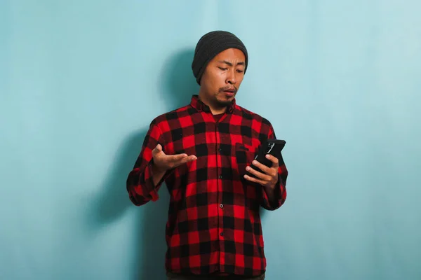 stock image An upset Young Asian man with a beanie hat and a red plaid flannel shirt is holding a mobile phone, experiencing problems, and feeling angry and annoyed, isolated on a blue background