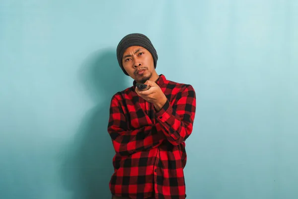 Bored young Asian man with a beanie hat and red plaid flannel shirt holding a remote control and looking bored while watching TV, isolated on a blue background