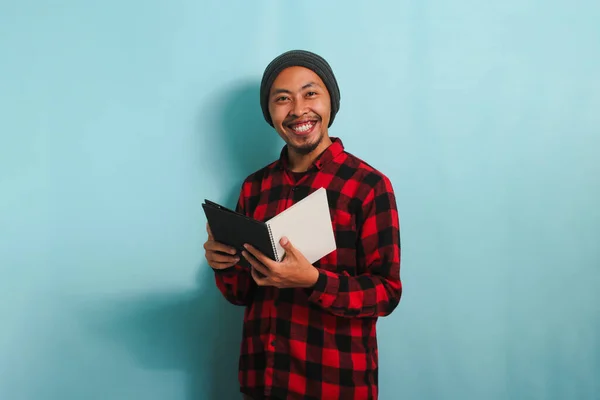 stock image A smiling young Asian man with a beanie hat and a red plaid flannel shirt is holding a book while standing against a blue background