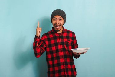 Şapkalı, kırmızı flanel gömlekli heyecanlı Asyalı genç adam elinde boş bir beyaz tabak tutarken gülümsüyor. Mavi bir arka planda ne yiyeceğine dair bir fikri var.