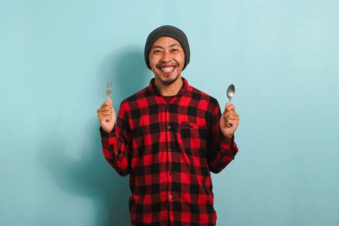 Şapkalı, kırmızı flanel gömlekli, elinde çatal ve kaşık tutan Asyalı genç adam, mavi bir arka planda izole edilmiş, mutlu ve yemek için istekli görünüyor.