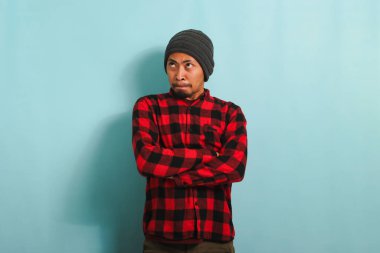 Şapkalı, kırmızı ekose gömlekli öfkeli bir Asyalı adam kollarını katlamış, rahatsız edici ve onaylamayan bir ifadeyle, mavi bir arka planda izole edilmiş bir şekilde açıklamalarını bekliyor.