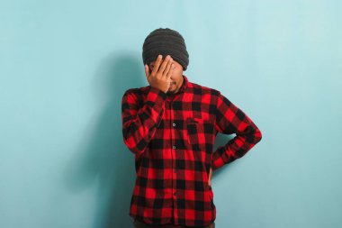 Utanmış ve utanmış bir Asyalı adam bereli ve kırmızı flanel gömlekli. Yüzünü mavi bir arka planda dururken eliyle örtüyor.