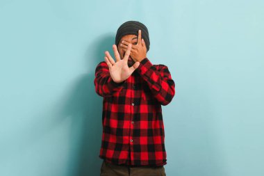 Şapkalı, kırmızı ekose gömlekli, korkmuş Asyalı genç adam avuçlarıyla gözlerini kapatıyor ve mavi bir arka planda gözlerden uzak bir şekilde parmaklarından süzülüyor.