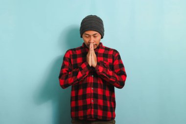 Bere şapkalı, kırmızı flanel gömlekli genç bir Asyalı Müslüman Müslüman, Müslüman Tanrı Allah 'a dua ediyor. Gözlerini mavi bir arka planda izole bir şekilde kapalı tut.