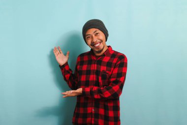 Heyecanlı, bereli ve kırmızı flanel gömlekli Asyalı genç bir adam elleri yanda görünüyor. Mavi arka planda izole edilmiş boş fotokopi alanını gösteriyor.