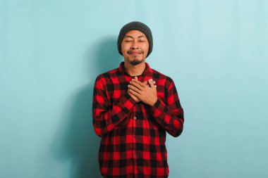 Şapkalı, kırmızı flanel gömlekli, minnettar, genç bir Asyalı ellerini kalbinde tutuyor. Rahatlamış ve müteşekkir. Mavi arka planda soyutlanmış.