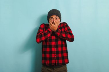 Şapkalı ve kırmızı flanel gömlekli genç Asyalı adam ağzını kapatıyor. Bu da demek oluyor ki, çenesini kapatmaya zorlanıyor. Mavi arka planda tek başına.