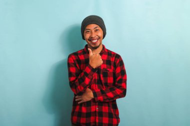 Şapkalı, kırmızı ekose gömlekli, kendine güvenen Asyalı genç adam iyi bir jest yapıyor ve kendini mavi arka planda yalnız hissediyor.