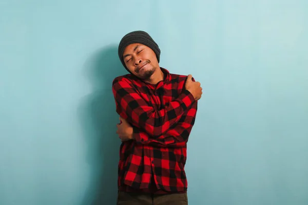 Young Asian man with beanie hat and red plaid flannel shirt hugs himself, expressing happiness and positivity, while standing against a blue background. Self love and self care concept.