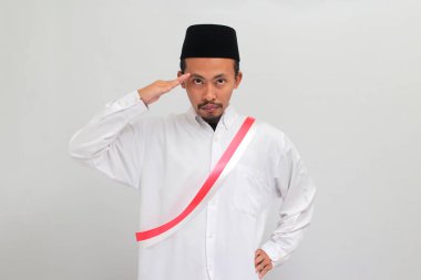 Endonezya 'nın heyecanlı genç adamı, kırmızı ve beyaz kuşak takıyor ve ya peci ya da kopiah, 17 Ağustos' ta Endonezya 'nın Bağımsızlık Günü' nü beyaz arka planda tek başına kutlarken selam veriyor.