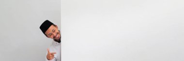 Endonezyalı heyecanlı genç bir adam, bir Songkok, peci veya kopiah giyiyor, beyaz boş bir afişin arkasında duruyor ve beyaz arka planda izole edilmiş bir reklam panosundaki boş bir fotokopi alanını işaret ediyor.