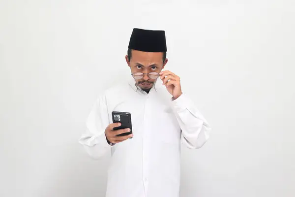 怒っている若いインドネシア人男性は ソングコックやピッチ またはコピアを着て 白い背景に隔離された携帯電話を保持しながら カメラを見下ろす眼鏡を見ています — ストック写真