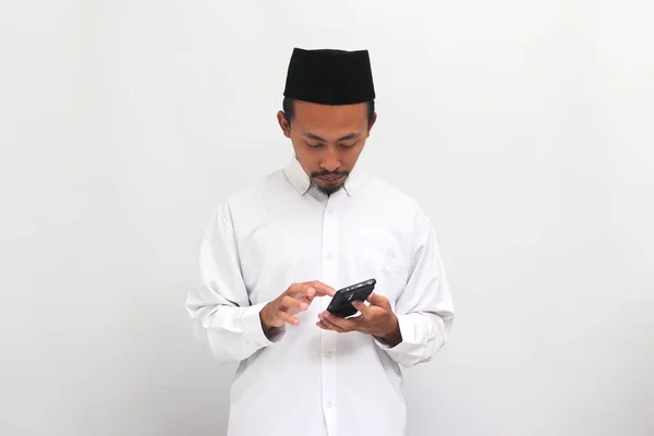 若いインドネシア人男性は ソングコック ピッチ またはコピアを着て 白い背景で孤立したソーシャルメディアをテキストまたはスクロールします — ストック写真