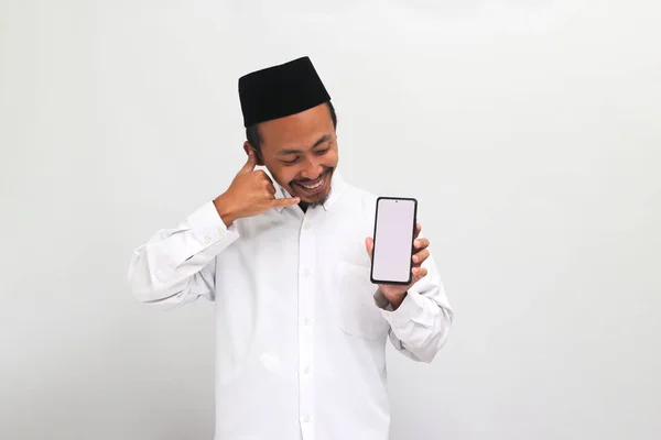 興奮した若いインドネシア人男性は ソングコックまたはピシアまたはピシアを着用し 彼の耳の近くに手を保持し 私をジェスチャーと呼んで 白い背景に隔離された白い画面の携帯電話を示しています — ストック写真