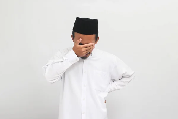 イライラした若いインドネシア人男性は ソングコック ピッチ またはコピアを身に着けているが 顔を覆い 白い背景に隔離された何か悪いことをしたことを後悔していると感じている — ストック写真