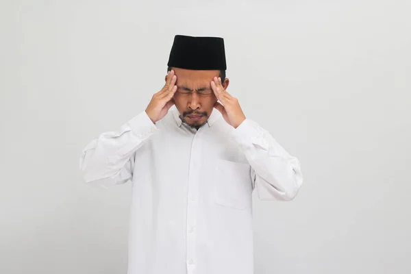 若いインドネシア人男性は ソングコック ピッチ またはコピアを着用して 頭痛に苦しんでおり 白い背景に隔離された痛みと片頭痛のために絶望的でストレスを感じています — ストック写真
