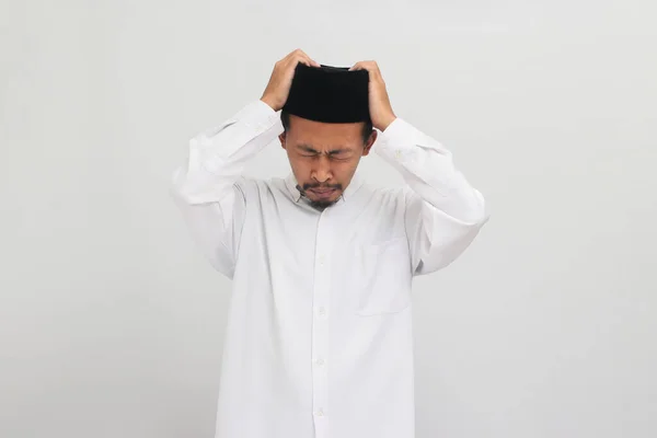 若いインドネシア人男性は ソングコック ピッチ またはコピアを着用して 頭痛に苦しんでおり 白い背景に隔離された痛みと片頭痛のために絶望的でストレスを感じています — ストック写真