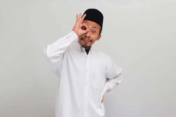 エキサイティングな若いインドネシア人男性は ソングコック ピッチ またはコピアを着用してOkのサインジェスチャーを指で行い 白い背景に隔離された彼の目で双眼鏡として働きます — ストック写真
