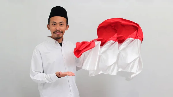 若いインドネシア人男性は ソングコック ピッチ またはコピアを着て 誇らしげに彼の横にインドネシアの旗を表示し 白い背景に孤立しています — ストック写真