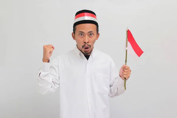 ワクワクする若いインドネシア人男性は ソングコック ピッチ またはコピアを着て 8月17日にインドネシアの独立記念日を祝い 白い背景に孤立した小さなインドネシアの旗を掲げています — ストック写真