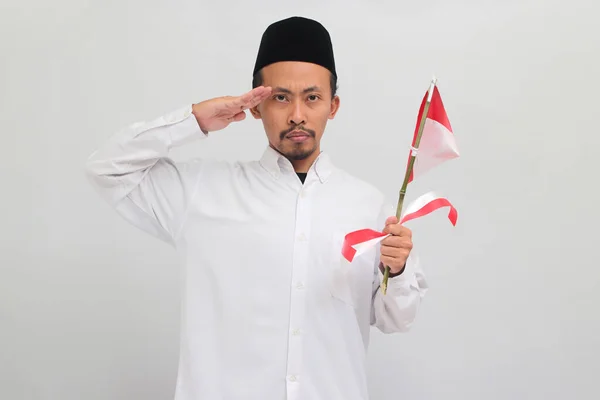 エキサイティングな若いインドネシア人男性は ソングコックを着て ピシア またはピシアを着て 8月17日に独立記念日を祝うために小さなインドネシアの旗を保持しながら敬礼のジェスチャーを作っています — ストック写真