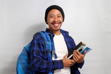 Kendine güvenen genç Asyalı bir öğrenci, bereli bir şapka ve gündelik bir gömlek giyer ve sırt çantası taşır. Elinde bir yığın kitap tutarken ve beyaz bir arka plan karşısında dururken mutlu görünür.