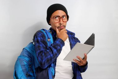 Düşünceli Asyalı bir öğrenci, gözlük takıyor, bere takıyor, gündelik giysiler giyiyor, sırt çantası taşıyor, beyaz bir arka plana yaslanıyor, bir kitap tutuyor ve elini düşünceli bir şekilde çenesine koyuyor.