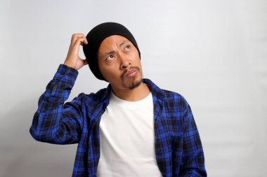 Kafası karışmış genç Asyalı bir adam, bereli bir şapka ve günlük giysiler içinde, bir çözüm ararken kafasını kaşıyor, şüphe ve kafa karışıklığını tasvir ediyor, beyaz bir arka plana karşı duruyor.